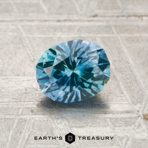 A 1.23-carat heated Montana sapphire is displayed on a textured light surface with the text "EARTH'S TREASURY" and a logo at the bottom.