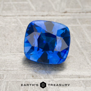 A 1.57-carat cushion-cut blue sapphire rests on a textured surface, with the Earth’s Treasury logo displayed at the bottom of the image.