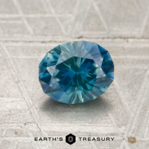 A 1.21-carat heated Montana sapphire, faceted in an oval shape with a blue-green hue, displayed on a textured gray surface featuring the text "Earth's Treasury" at the bottom.