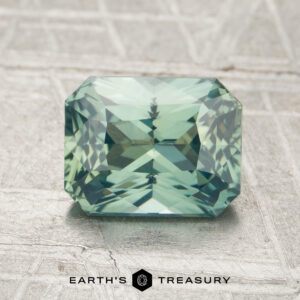 A 1.56-carat rectangular, greenish-blue faceted sapphire rests on a textured surface with the "Earth's Treasury" logo and text below.