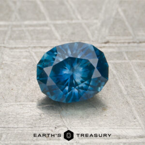 A 1.13-carat heated Montana sapphire in an oval cut is displayed on a textured surface, with the "Earth's Treasury" logo and text at the bottom.
