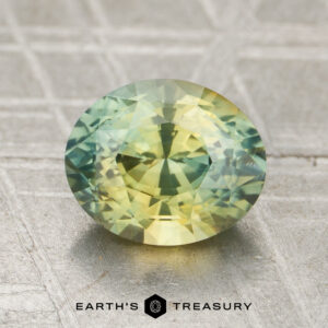 A 1.52-carat oval-shaped faceted sapphire featuring yellow and green hues is displayed on a textured metallic surface, with the "Earth's Treasury" logo at the bottom.