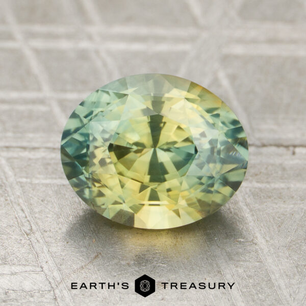 1.52-Carat Sapphire A 1.52-carat oval-shaped faceted sapphire featuring yellow and green hues is displayed on a textured metallic surface, with the "Earth's Treasury" logo at the bottom.