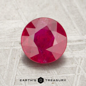 A 1.30-carat round, faceted ruby is displayed on a gray textured surface with the words "EARTH'S TREASURY" below it.