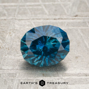 A 1.70-carat heated Montana sapphire is displayed on a textured gray surface with "EARTH'S TREASURY" at the bottom.