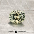 A round, faceted pale yellow-green 1.02-carat heated Montana sapphire rests on a textured metallic surface, with the text "EARTH'S TREASURY" at the bottom.