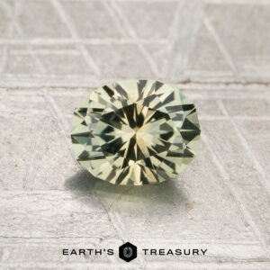 A round, faceted pale yellow-green 1.02-carat heated Montana sapphire rests on a textured metallic surface, with the text "EARTH'S TREASURY" at the bottom.