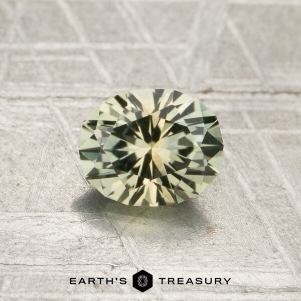 1.02-Carat Mint Green-Yellow Particolored Montana Sapphire (Heated) A round, faceted pale yellow-green 1.02-carat heated Montana sapphire rests on a textured metallic surface, with the text "EARTH'S TREASURY" at the bottom.