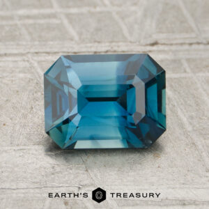 A 1.17-carat rectangular blue-green sapphire with emerald cut facets is displayed on a textured grey surface, with the "Earth's Treasury" logo at the bottom.