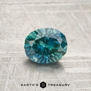 A 0.96-carat heated Montana sapphire in a round, faceted cut is shown on a textured gray surface with the "Earth's Treasury" logo below.