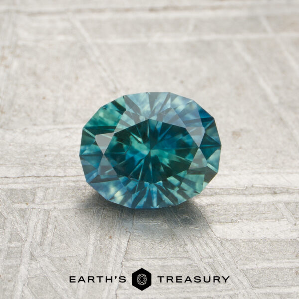 0.96-Carat Teal Blue-Green Particolored Montana Sapphire (Heated) A 0.96-carat heated Montana sapphire in a round, faceted cut is shown on a textured gray surface with the "Earth's Treasury" logo below.