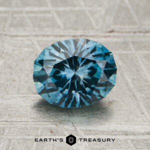 A 1.51-carat heated Montana sapphire is displayed on a textured gray surface, with "EARTH'S TREASURY" and a geometric logo shown below the gemstone.