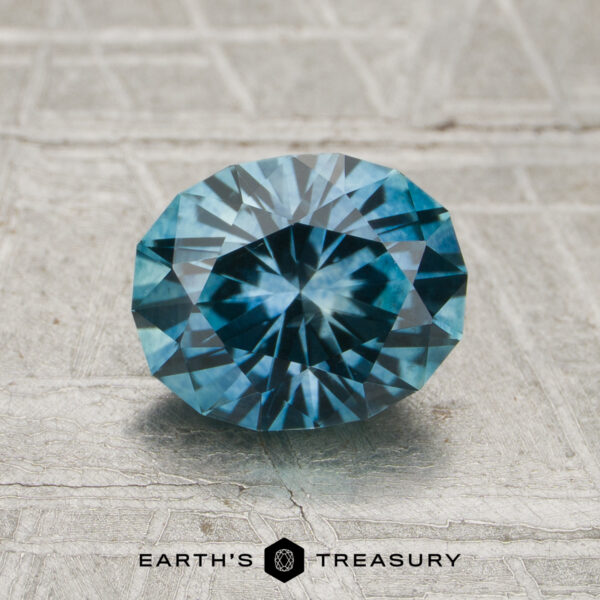 1.51-Carat Teal Montana Sapphire (Heated) A 1.51-carat heated Montana sapphire is displayed on a textured gray surface, with "EARTH'S TREASURY" and a geometric logo shown below the gemstone.
