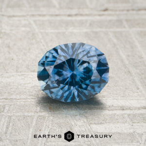 A 1.08-carat heated Montana sapphire in an oval cut is displayed on a textured gray surface, with the "Earth's Treasury" logo and text shown below.