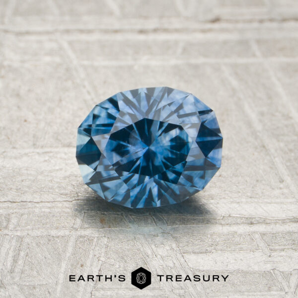 A 1.08-carat heated Montana sapphire in an oval cut is displayed on a textured gray surface, with the "Earth's Treasury" logo and text shown below.