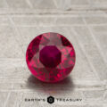 A 1.18-carat round-cut ruby rests on a textured surface, with the "EARTH'S TREASURY" logo and text visible at the bottom of the image.