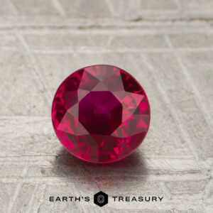 A 1.18-carat round-cut ruby rests on a textured surface, with the "EARTH'S TREASURY" logo and text visible at the bottom of the image.