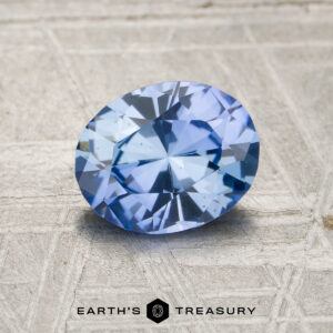 A 1.53-carat unheated Ceylon sapphire, oval-cut and light blue, rests on a textured surface with the "Earth's Treasury" logo and text at the bottom of the image.