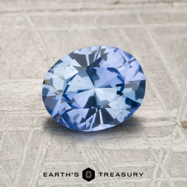 A 1.53-carat unheated Ceylon sapphire, oval-cut and light blue, rests on a textured surface with the "Earth's Treasury" logo and text at the bottom of the image.