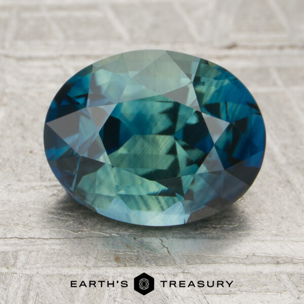 2.72-Carat Sapphire (Heated) A 2.72-carat heated sapphire, oval-cut and blue-green in color, rests on a textured surface featuring the "Earth's Treasury" name and logo below the gem.