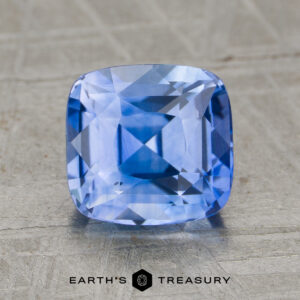A 2.60-carat heated sapphire, cushion-cut and blue, is displayed on a textured surface with "EARTH'S TREASURY" and a hexagonal logo beneath it.