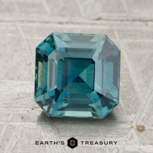 A 2.55-carat heated sapphire with an emerald cut and blue-green hue is displayed on a light textured surface, with the text "EARTH'S TREASURY" below.