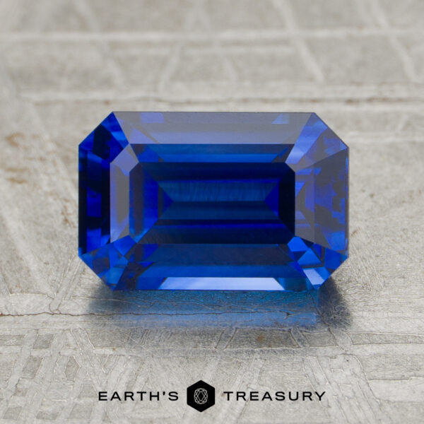 2.47-Carat Sapphire (Heated A 2.47-carat heated sapphire, emerald-cut and rectangular, is shown on a textured gray surface with the "EARTH'S TREASURY" logo at the bottom.