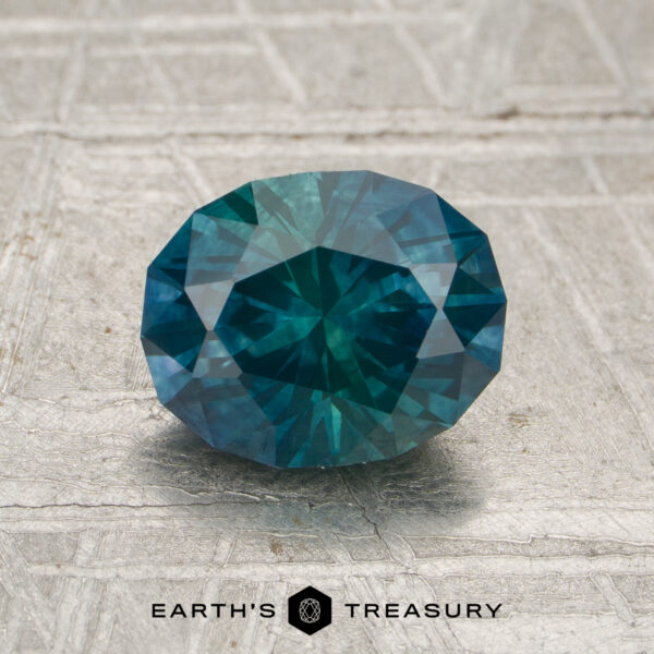 1.68-Montana Teal-Green Particolored Sapphire (Heated) A 1.68-carat heated Montana Sapphire with an oval, faceted cut and blue-green hue is displayed on a textured surface, with the "Earth's Treasury" name and logo beneath.