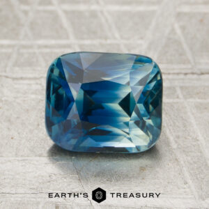 A 2.32-carat heated sapphire with a cushion cut rests on a textured metallic surface, with "Earth's Treasury" logo and text displayed beneath.