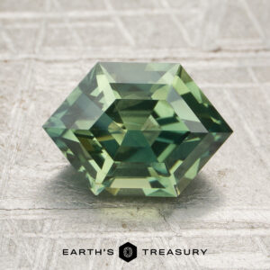 A 2.38-carat heated sapphire, cut in a green hexagonal shape with step facets, rests on a textured surface. The "EARTH'S TREASURY" logo and text appear at the bottom.