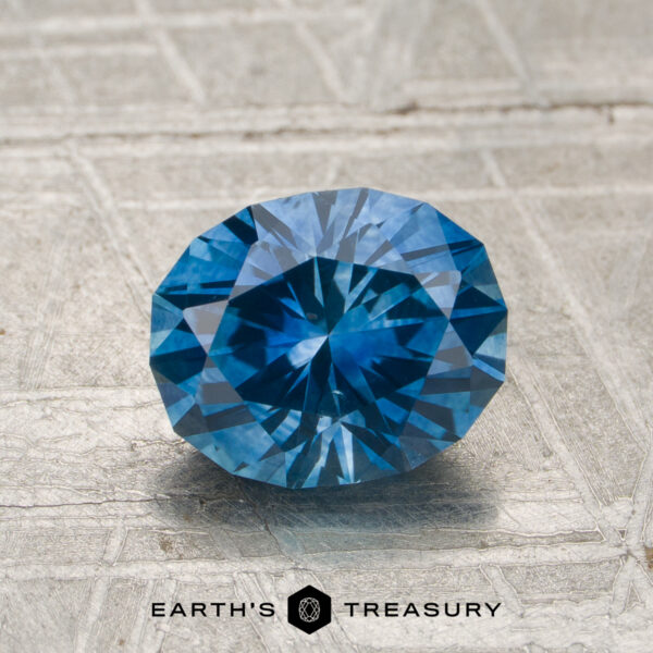 1.68-Carat Teal Blue Montana Sapphire (Heated) A 1.68-carat heated Montana sapphire is showcased on a textured surface, with the "Earth's Treasury" logo and text visible at the bottom.