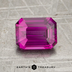Rectangular emerald-cut 2.05-carat heated sapphire in bright pink, displayed on a textured metallic surface with the text "Earth's Treasury" at the bottom.