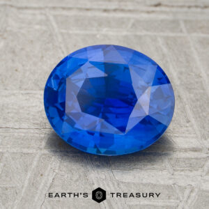 A 2.03-carat heated blue sapphire with an oval cut rests on a textured gray surface, displayed above the Earth's Treasury logo and name.