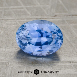 2.02-carat heated sapphire, oval-cut and blue, displayed on a textured gray surface with "EARTH'S TREASURY" text and logo beneath.