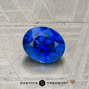 A 1.25-carat heated sapphire, oval-cut and blue, sits on a textured metallic surface with "EARTH'S TREASURY" displayed below.