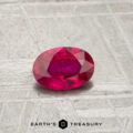 A 0.54-carat oval-cut ruby with a pinkish-red hue rests on a textured gray surface above the "Earth's Treasury" logo.