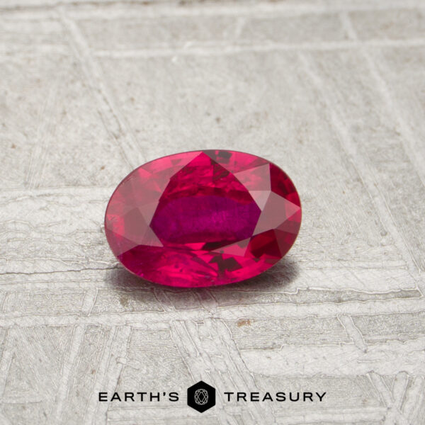 0.54-Carat Mozambique Ruby (Heated) A 0.54-carat oval-cut ruby with a pinkish-red hue rests on a textured gray surface above the "Earth's Treasury" logo.