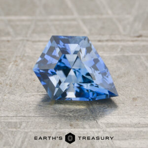 A faceted 1.13-carat heated blue sapphire with a unique geometric shape is displayed on a textured surface, with "Earth's Treasury" below.