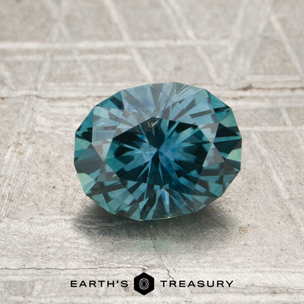 1.63-Carat Teal Montana Sapphire (Heated) A 1.63-carat heated Montana sapphire, cut in a faceted oval shape, is shown on a textured surface with the "Earth's Treasury" logo and text at the bottom.