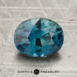 A 2.44-carat unheated sapphire with an oval cut and blue-green hue rests on a light textured surface, with “EARTH'S TREASURY” displayed at the bottom.