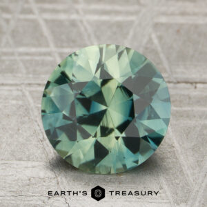 A round, faceted blue-green 2.40-carat unheated sapphire is displayed on a textured gray surface with the "Earth's Treasury" logo beneath it.