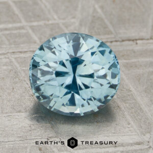 A 2.36-Carat Unheated Sapphire, faceted in an oval shape and light blue in color, rests on a textured surface with the "Earth's Treasury" name and hexagon logo visible below.