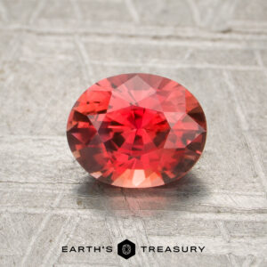 A 1.11-carat heated oval-cut sapphire with intricate facets rests on a textured gray surface, with "Earth's Treasury" displayed below.