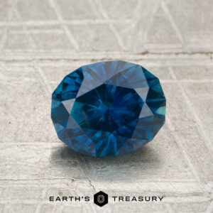 A 1.62-carat heated Montana sapphire with a round cut is displayed on a textured gray surface, featuring the "Earth's Treasury" logo and text at the bottom.
