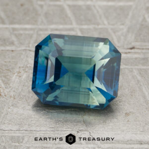 A 2.30-carat unheated sapphire, emerald-cut in blue-green hues, is shown on a metallic textured surface with the Earth’s Treasury logo at the bottom.