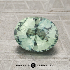 A 2.05-carat unheated sapphire with an oval cut and pale green hue is displayed on a textured gray surface featuring the Earth’s Treasury logo and text below.