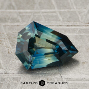A faceted, blue-green 2.04-carat heated sapphire is displayed on a textured gray surface, with the "EARTH'S TREASURY" logo and text at the bottom.