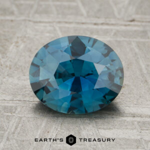 A 2.03-carat heated Madagascar sapphire with a faceted oval cut and blue-green hue is displayed on a textured surface, with "EARTH'S TREASURY" written below.