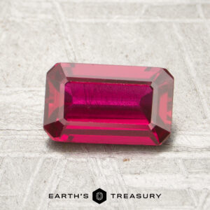 A 1.13-carat faceted ruby is shown on a textured surface with the "Earth’s Treasury" logo beneath it.