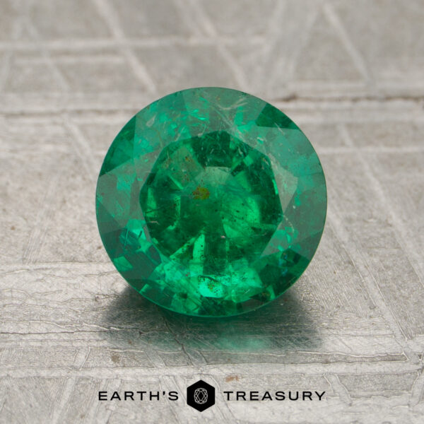 A 1.34-carat round-cut emerald gemstone displayed on a textured gray surface with "EARTH'S TREASURY" at the bottom.